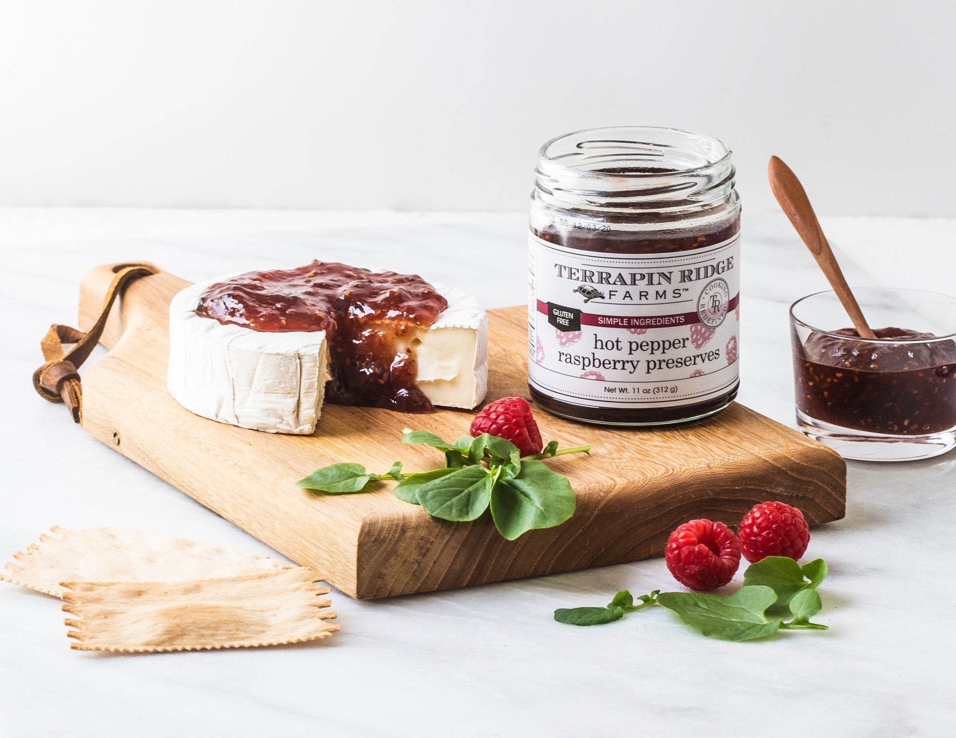 jar of Hot Pepper Raspberry Preserves set on a wooden board with a round of brie topped iwth Hot Pepper Raspberry Preserves, fresh raspberries, and herb leaves.