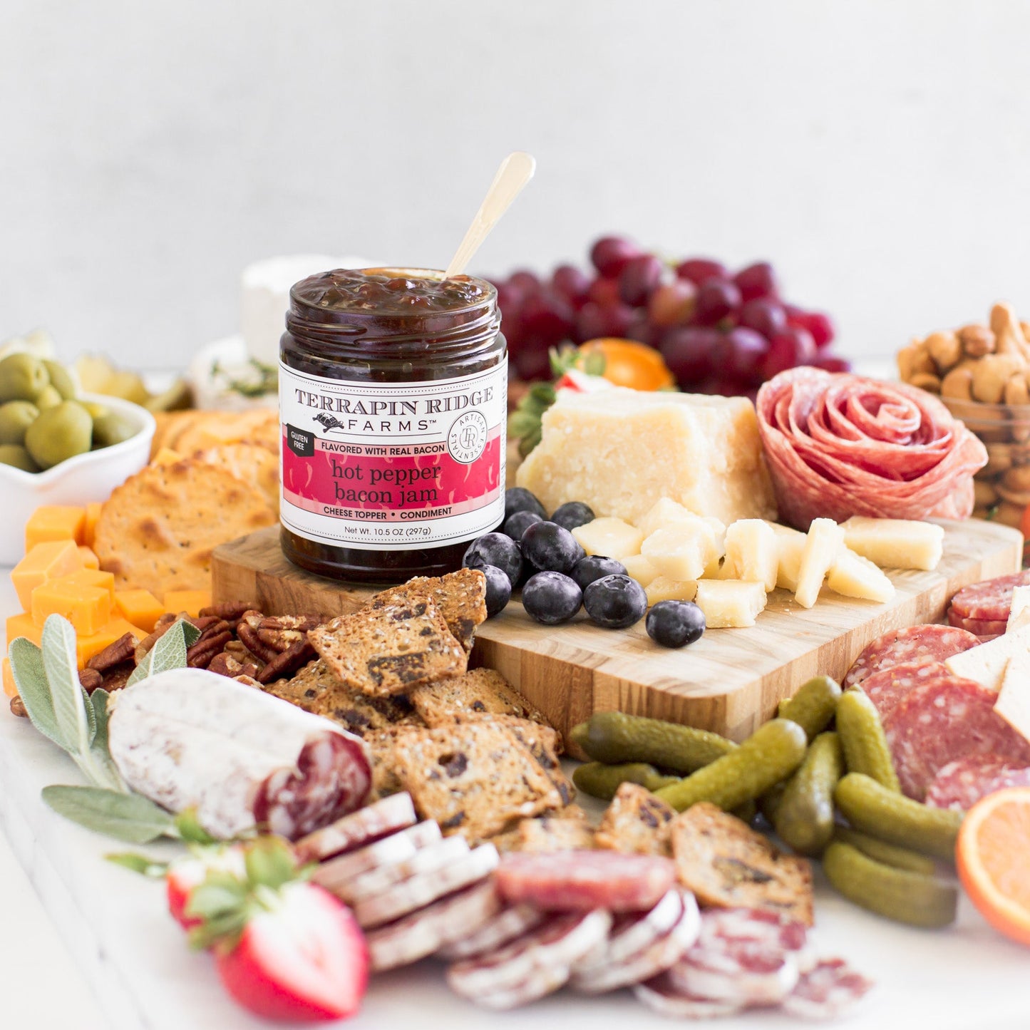 jar of Hot Pepper Bacon Jam on a board with meats, crackers, cheese, and fruits.