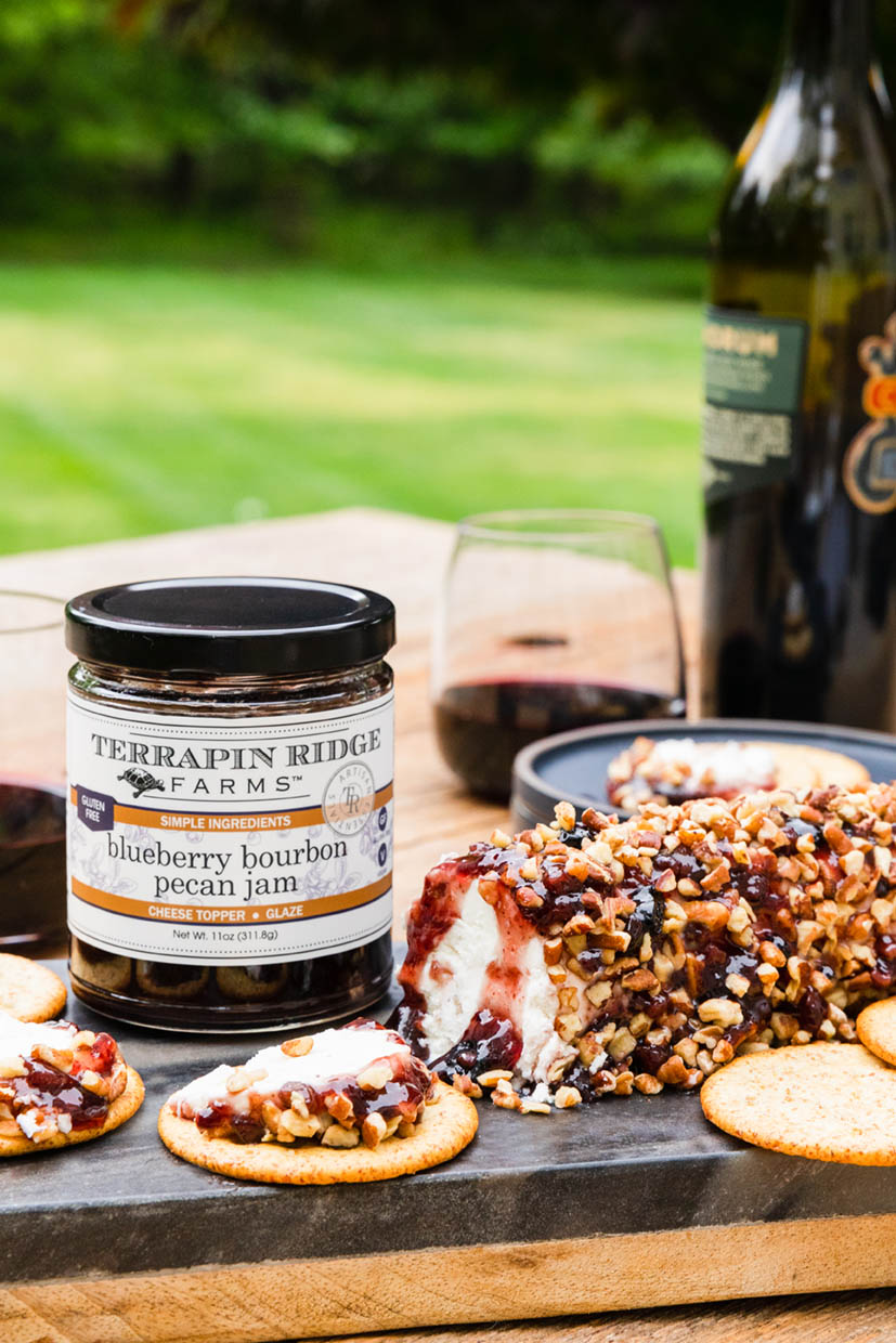 jar of Blueberry Bourbon Pecan Jam set on a table with appetizers, a bottle, and glasses of wine.
