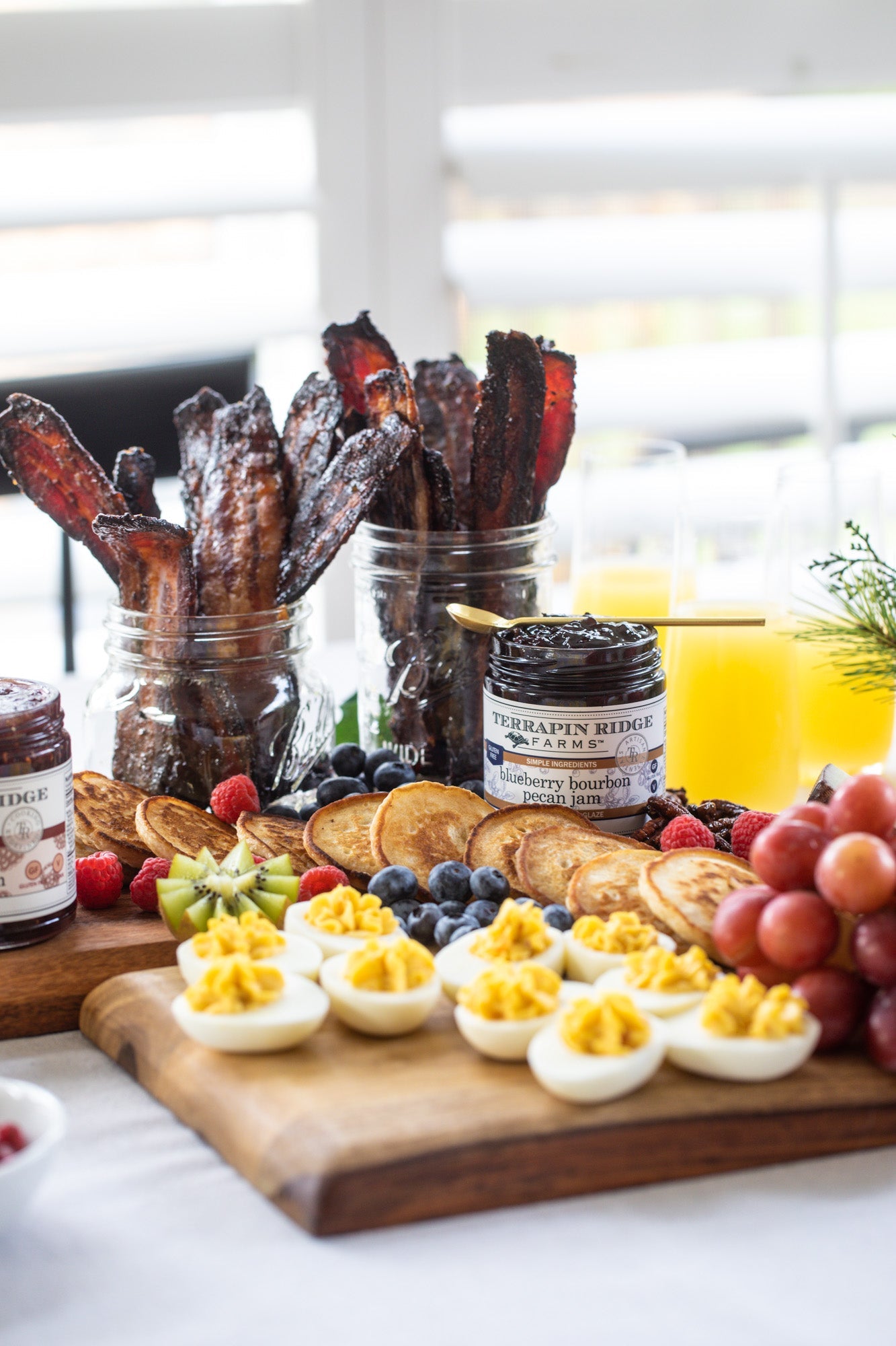jar of Blueberry Bourbon Pecan Jam set on a charcuterie board with eggs, pancakes, fruits, and bacon.