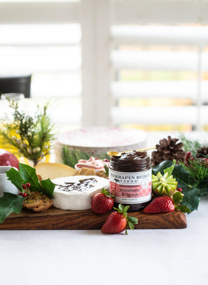 jar of strawberry fig jam set on a board with cheeses and fruit arranged with greenery