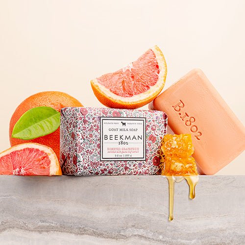 wrapped bar of Honeyed Grapefruit Goat Milk Bar Soap with an unwrapped bar of soap leaning against it arranged with grapefruit wedges and a honeycomb dripping with honey.