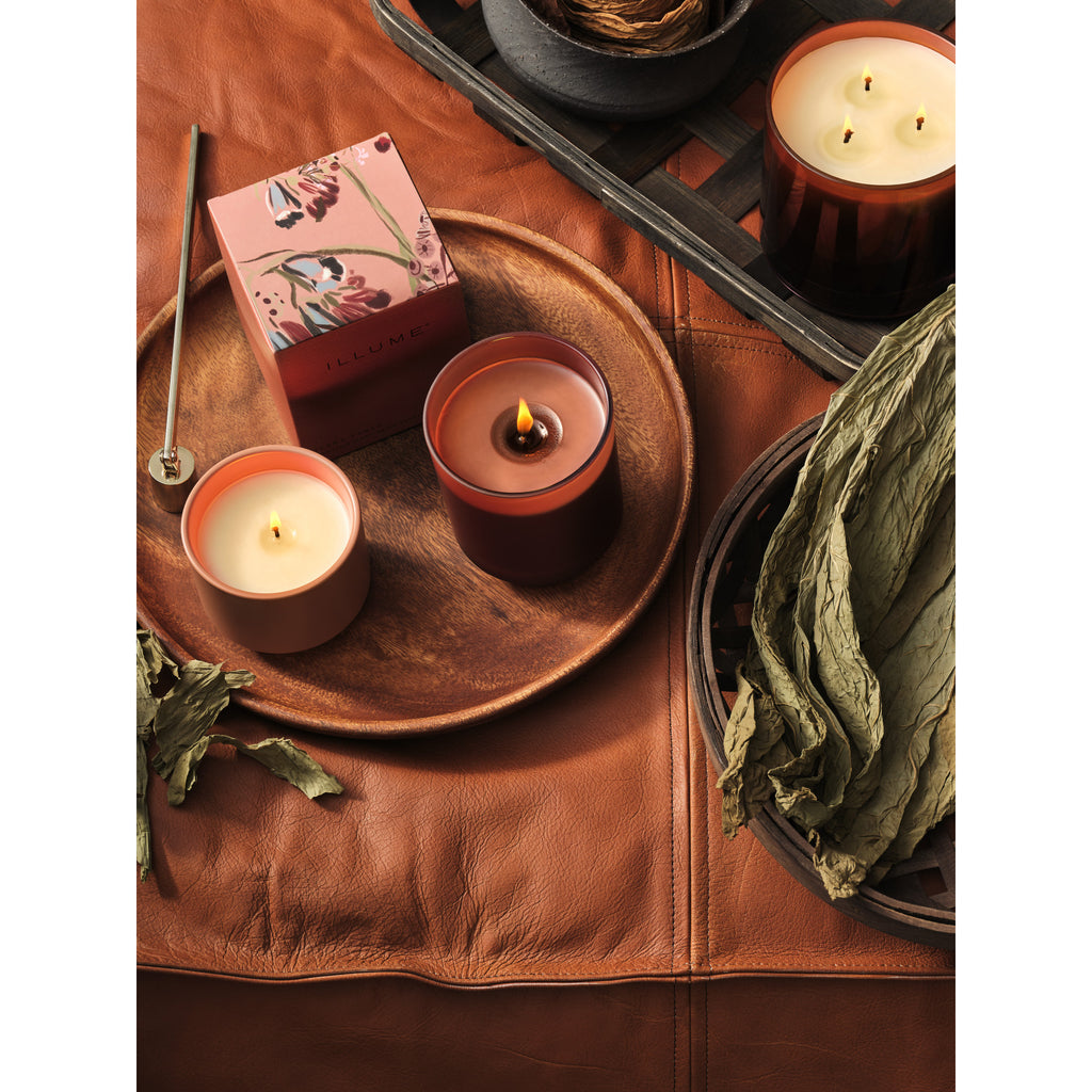 gold candle snuffer displayed on a round wooden tray next to two candles, and a colorful box on a leather ottoman