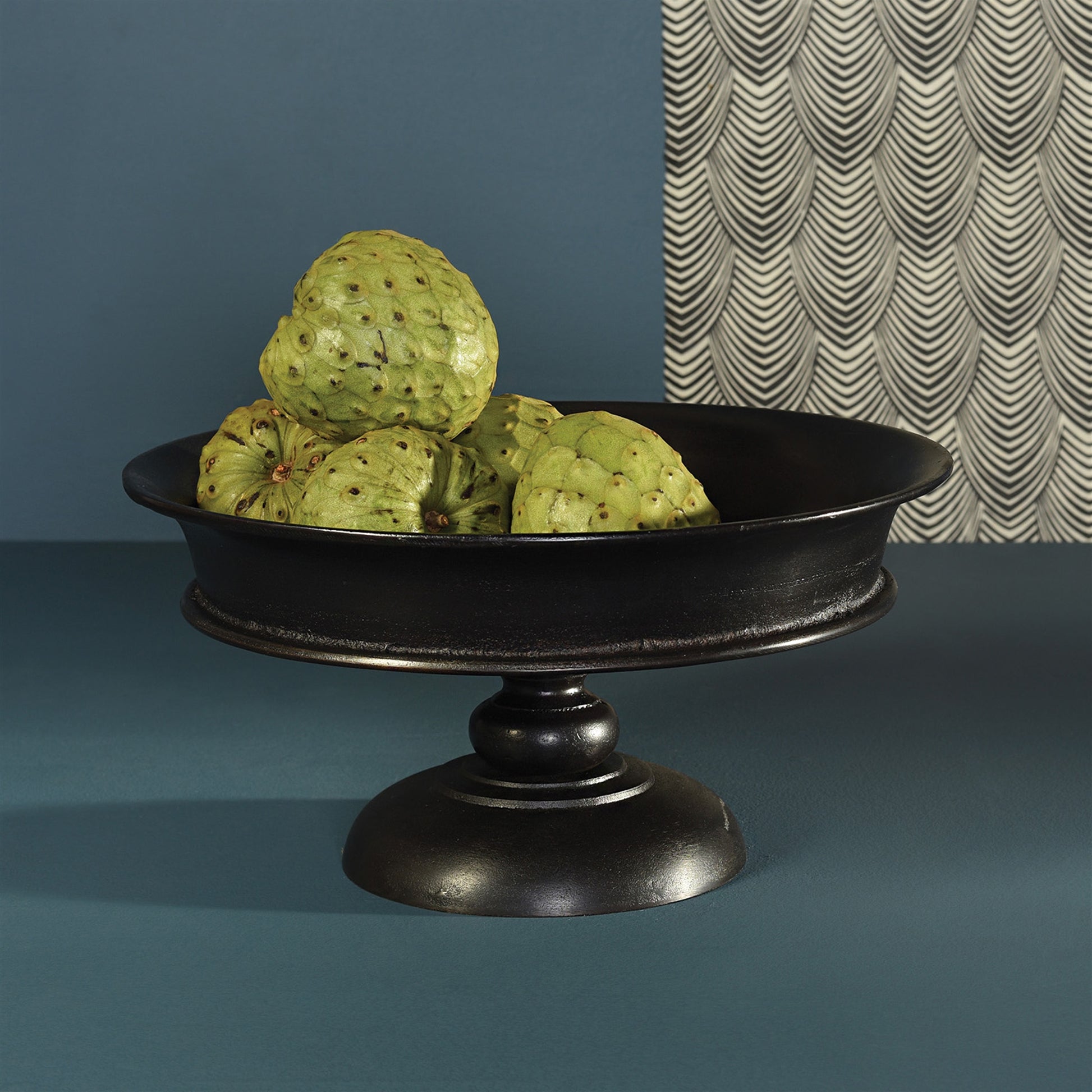 black metal pedestal bowl filled with green fruit set on a blue table.