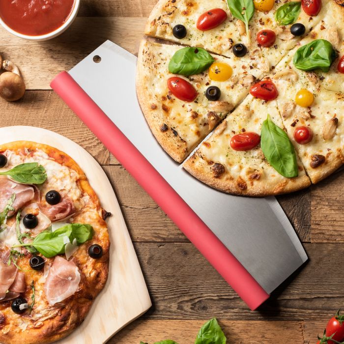 Two pizzas on a wooden table with a pizza cutter, surrounded by ingredients.