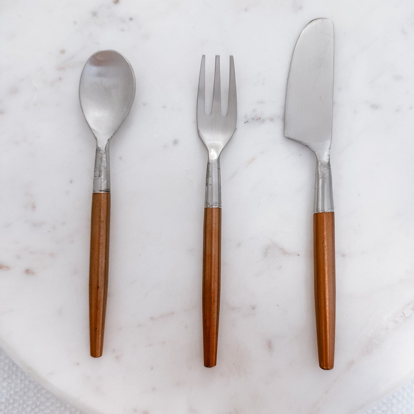 small spoon, fork, and spreader with copper handles in a row on a marble countertop.