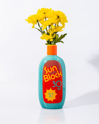 A ceramic vase designed to resemble a bottle of sunblock, with a bright label and an orange cap, now being used as a vase for yellow flowers.