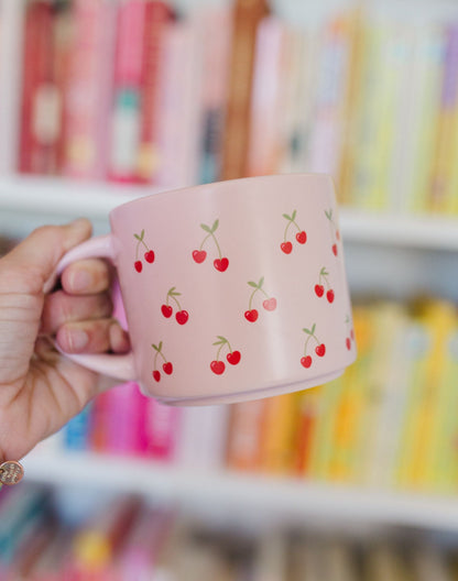 Hand holding a pink mug with cherry design against a blurred colorful background