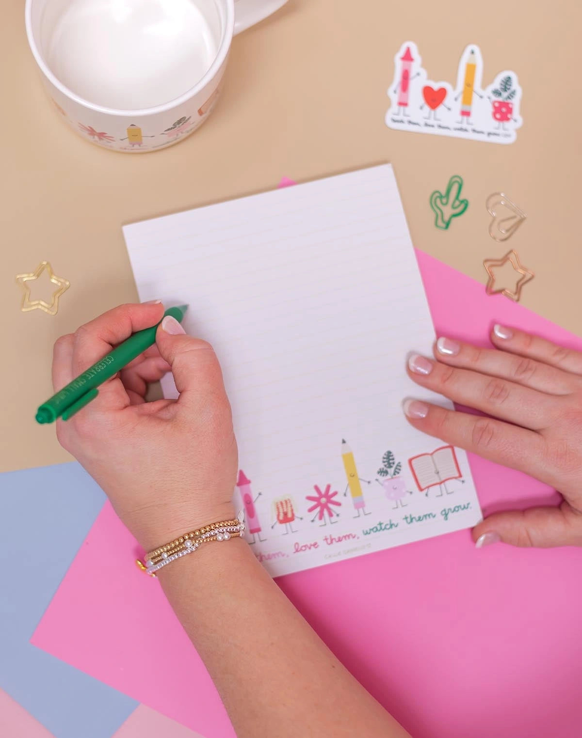 person's hands writing on a lined notepad with a green pen. The notepad features cute illustrations of school supplies and the phrase 'Love them, watch them, watch them grow'.