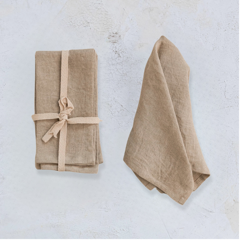 Two sets of stonewashed linen napkins on a plaster background. First set is a set of napkins tied with twill ribbon, second set is unfolded and laid next to the set.