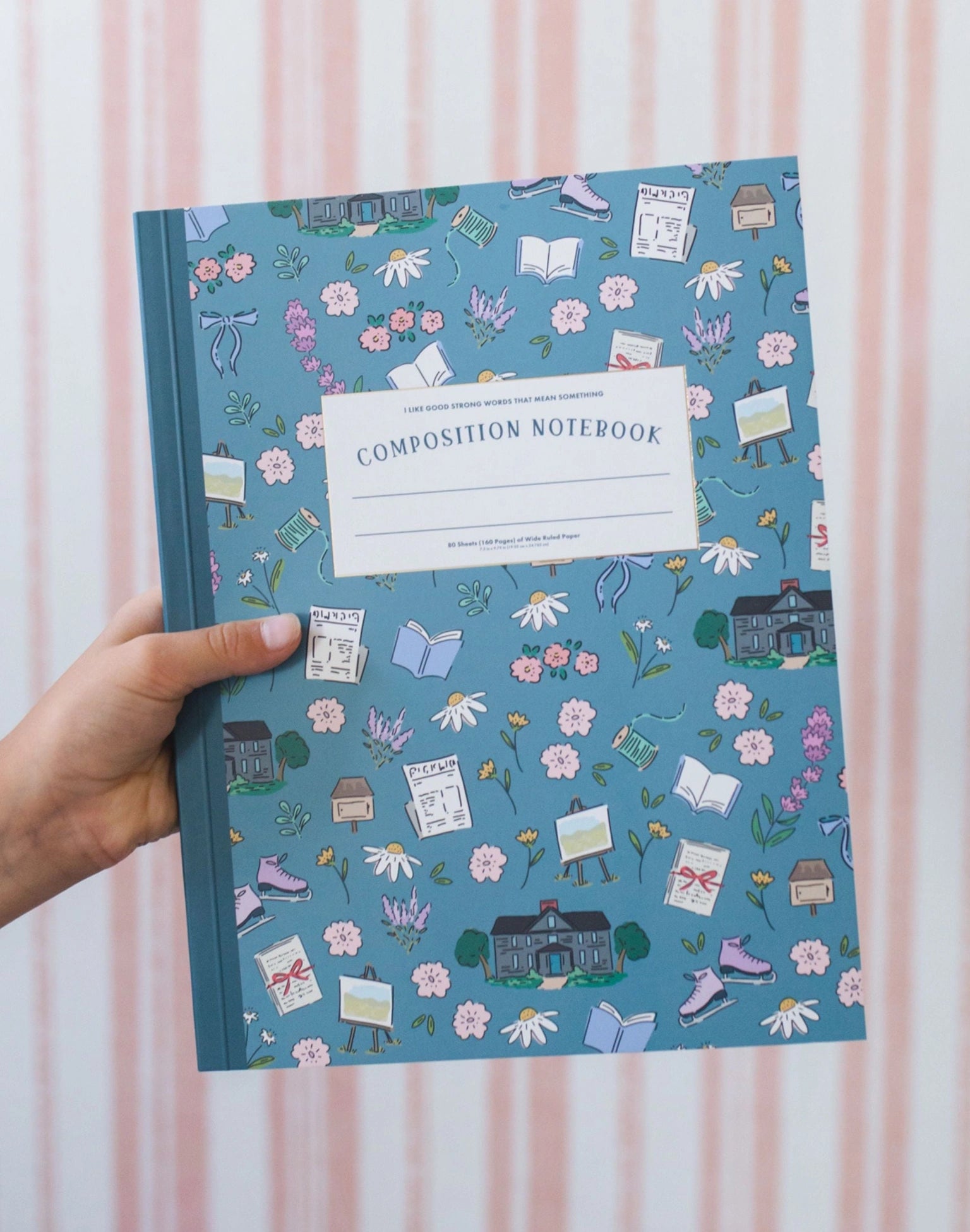 Hand holding a blue composition notebook with a decorative cover against a pink striped background