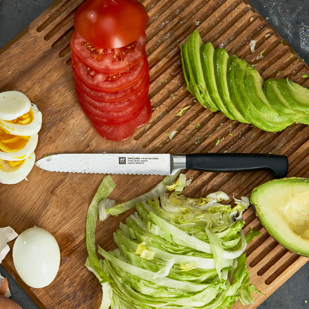 Four Star 5 Inch Serrated Utility Knife laying on a cutting board surrounded by sliced veggies and boiled eggs.