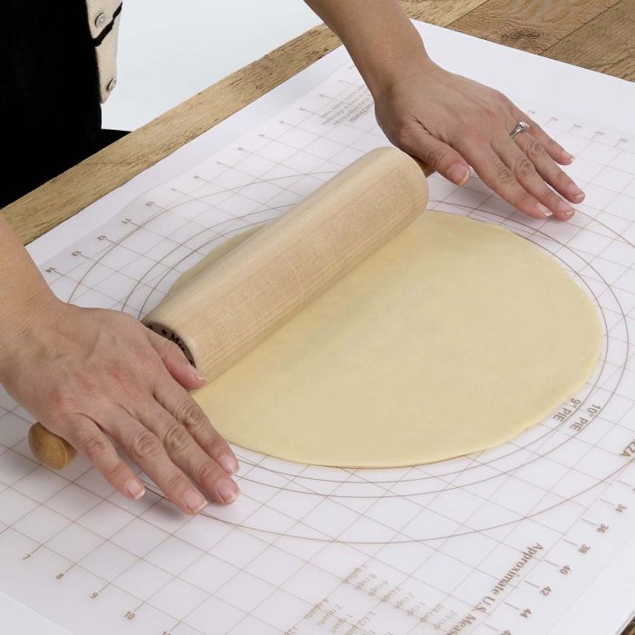 close-up of Person rolling out dough with a wooden rolling pin on a pastry mat.