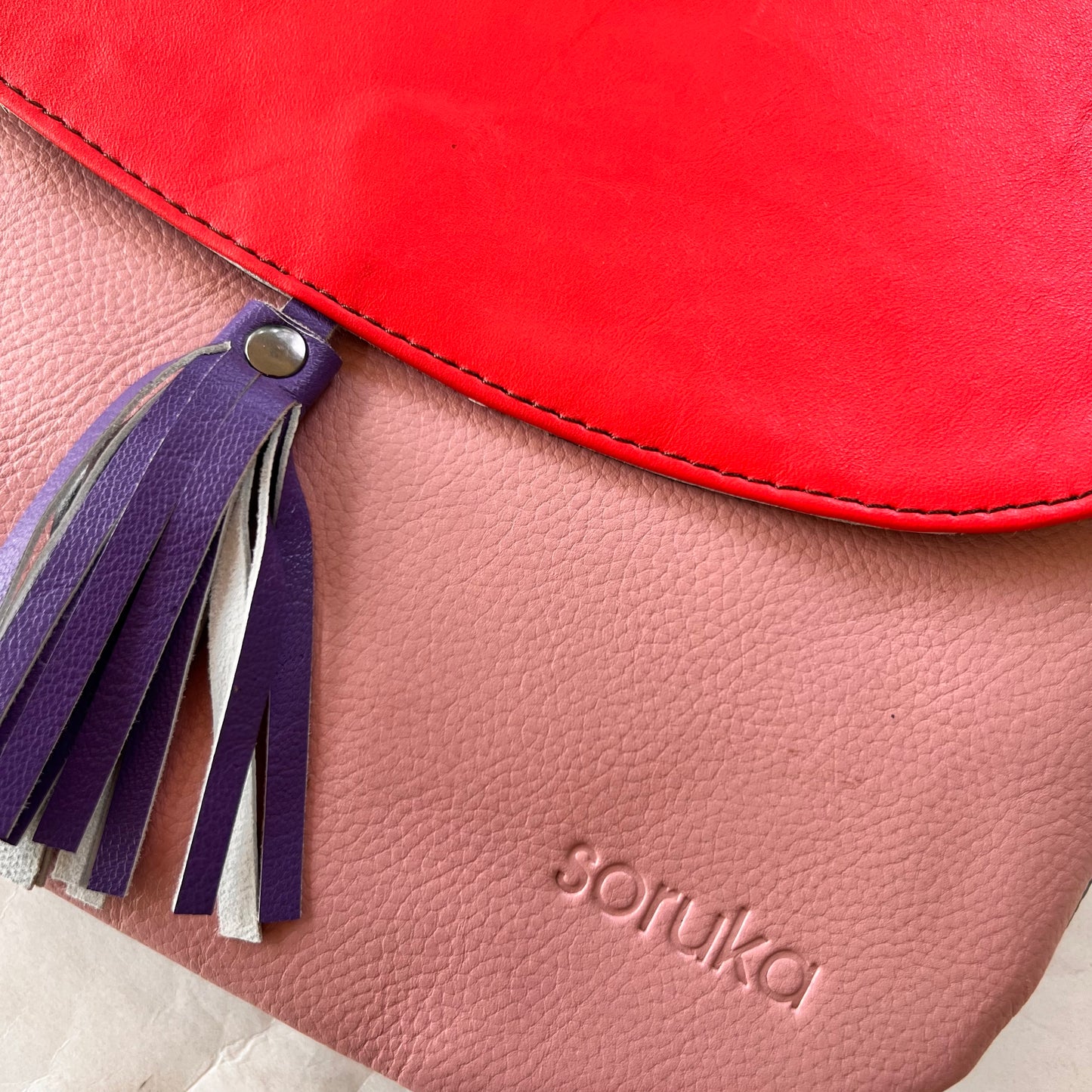 close-up of lola bag with bright red flap with purple tassel over a pink body.
