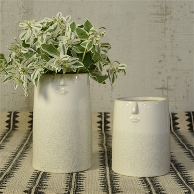 2 sizes of face vases set on a black and white patterned table, the larger vase is filled with greenery.