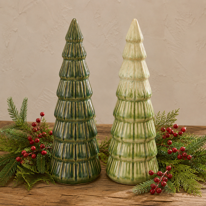 Two ceramic Christmas trees with a ribbed, tiered design, one dark green and one light green with cream highlights, displayed on a rustic wooden surface with green pine branches and red berries against a textured beige wall.