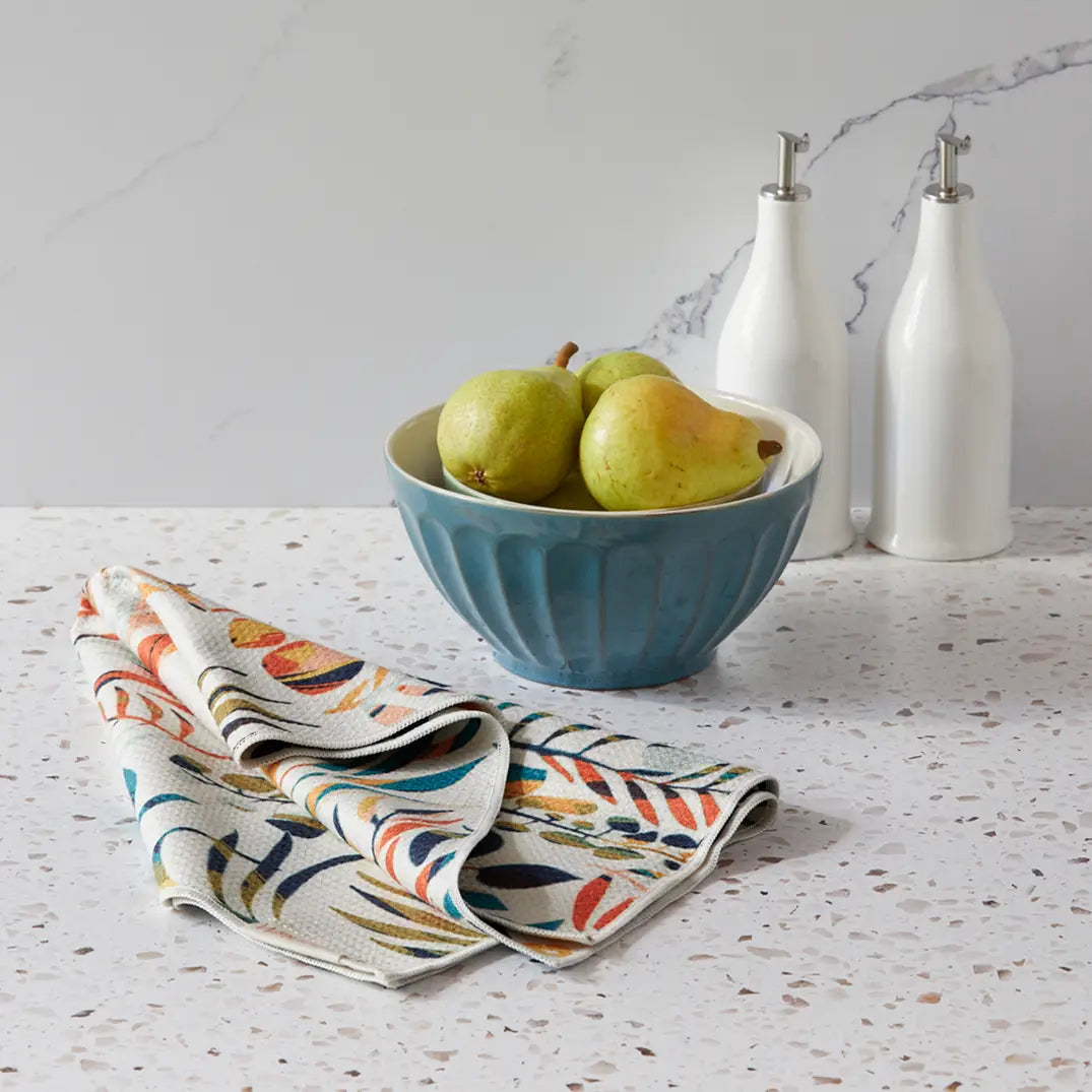 Colorful leaves dish towel draped showing the leaves side sitting on a counter next to a bowl full of pears.