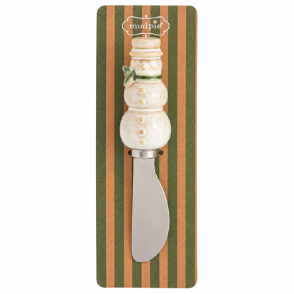 A snowman-handled spreader, featuring a cream-colored ceramic snowman handle with buttons and a green scarf, mounted on a green and tan striped cardboard backing.
