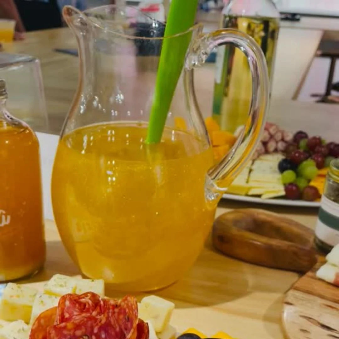 Clear pitcher filled with an orange glittery drink on a table with cheese and fruit platter.