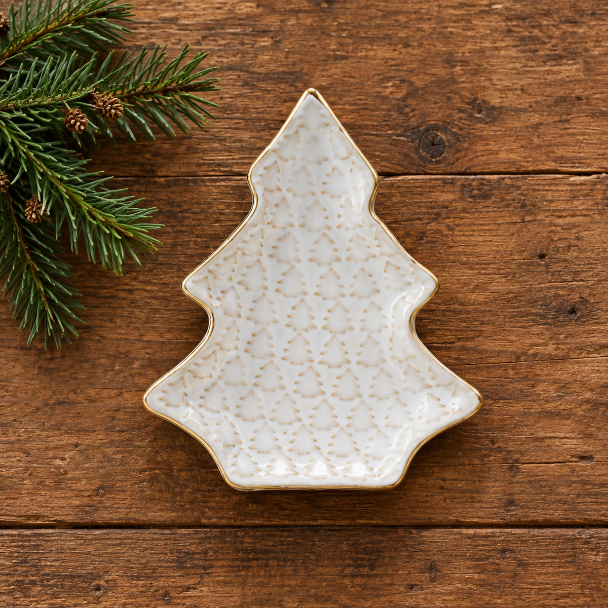 A Christmas tree-shaped ceramic dish with a textured cream glaze and gold trim, placed on a rustic wooden surface next to pine branches.