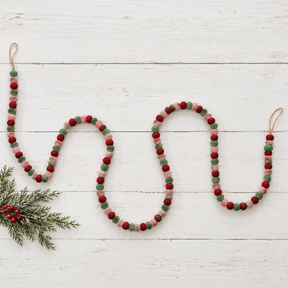 felted pom garland in alternating colors of red pink and green draped on white wooden surface with a sprig of greenery