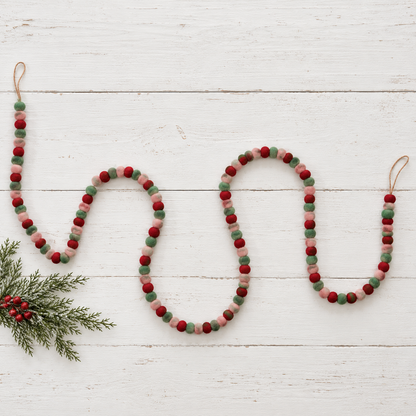 felted pom garland in alternating colors of red pink and green draped on white wooden surface with a sprig of greenery