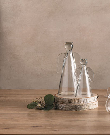 Decorative glass angel figures on a wooden surface with a sprig of greenery shown on a neutral background