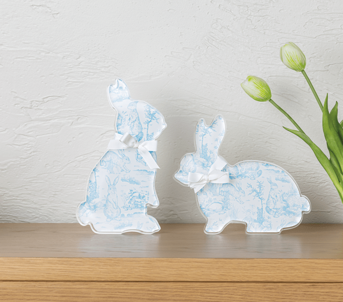 Two decorative acrylic bunny figurines with blue toile patterns on a wooden surface with tulips in the background.