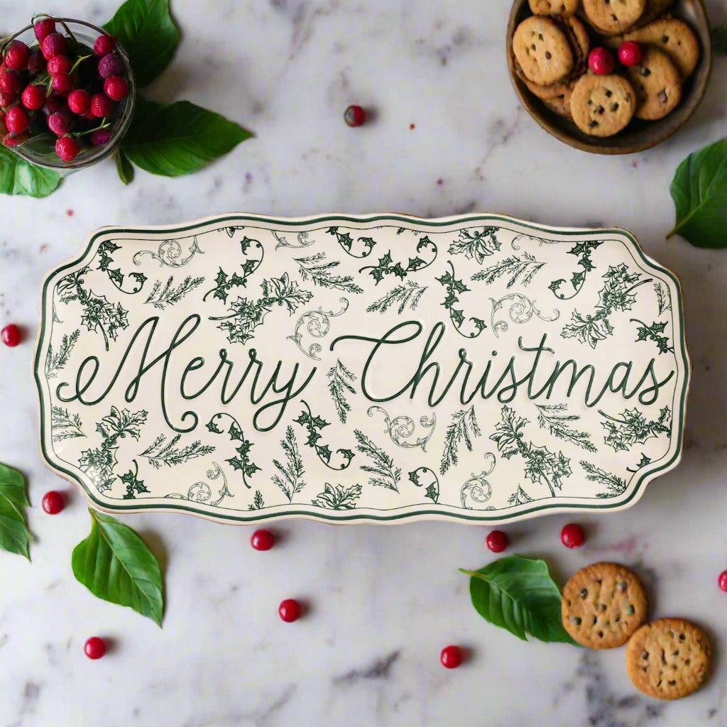 Decorative tray with 'Merry Christmas' text and green leaf patterns on a marble surface with red berries, cookies, and leaves around it