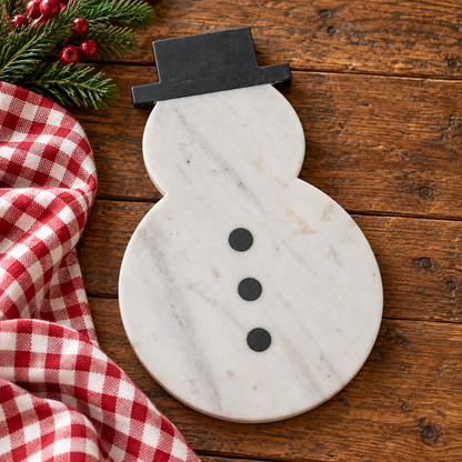 A snowman-shaped serving board made of white marble with gray veining, featuring a black hat silhouette and three black buttons. It is styled on a rustic wooden table next to a red and white checkered cloth and green pine branches with red berries.