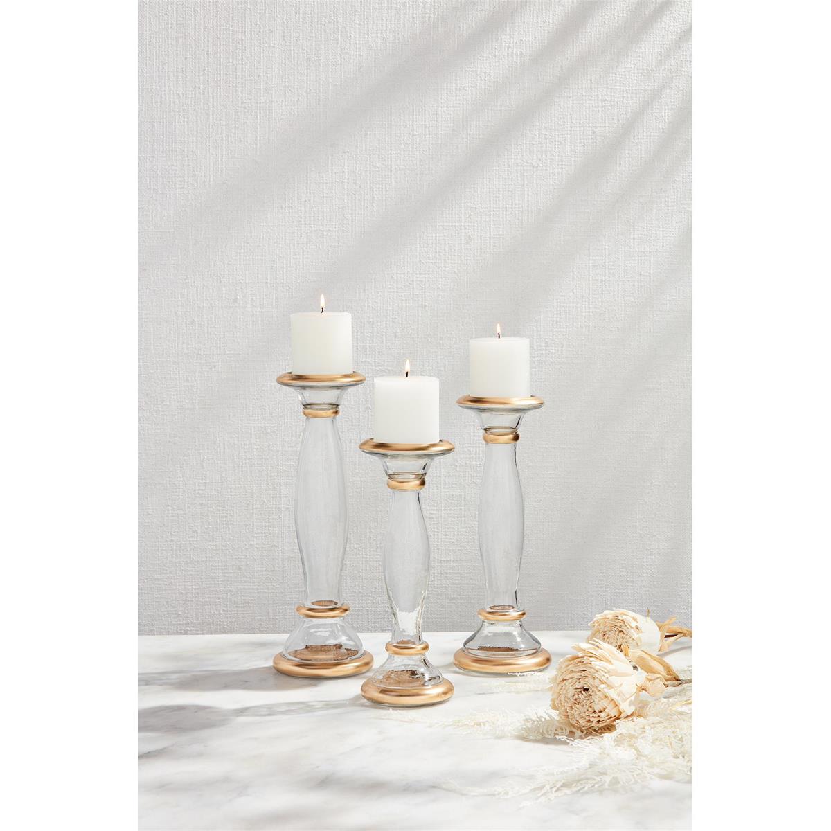 3 sizes of glass and gold candlesticks with lit candles on them set on a table with dried flowers.