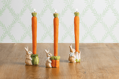 Three taper candle holders with rabbit figures on a wooden surface and patterned wall background.