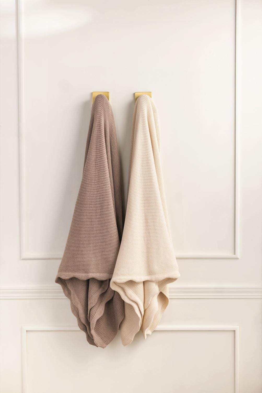Two beige knit blankets hanging on a white paneled wall.