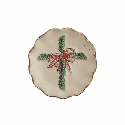 A scalloped ceramic plate with a hand-painted cross made of pine branches with red bow shown on a white background.