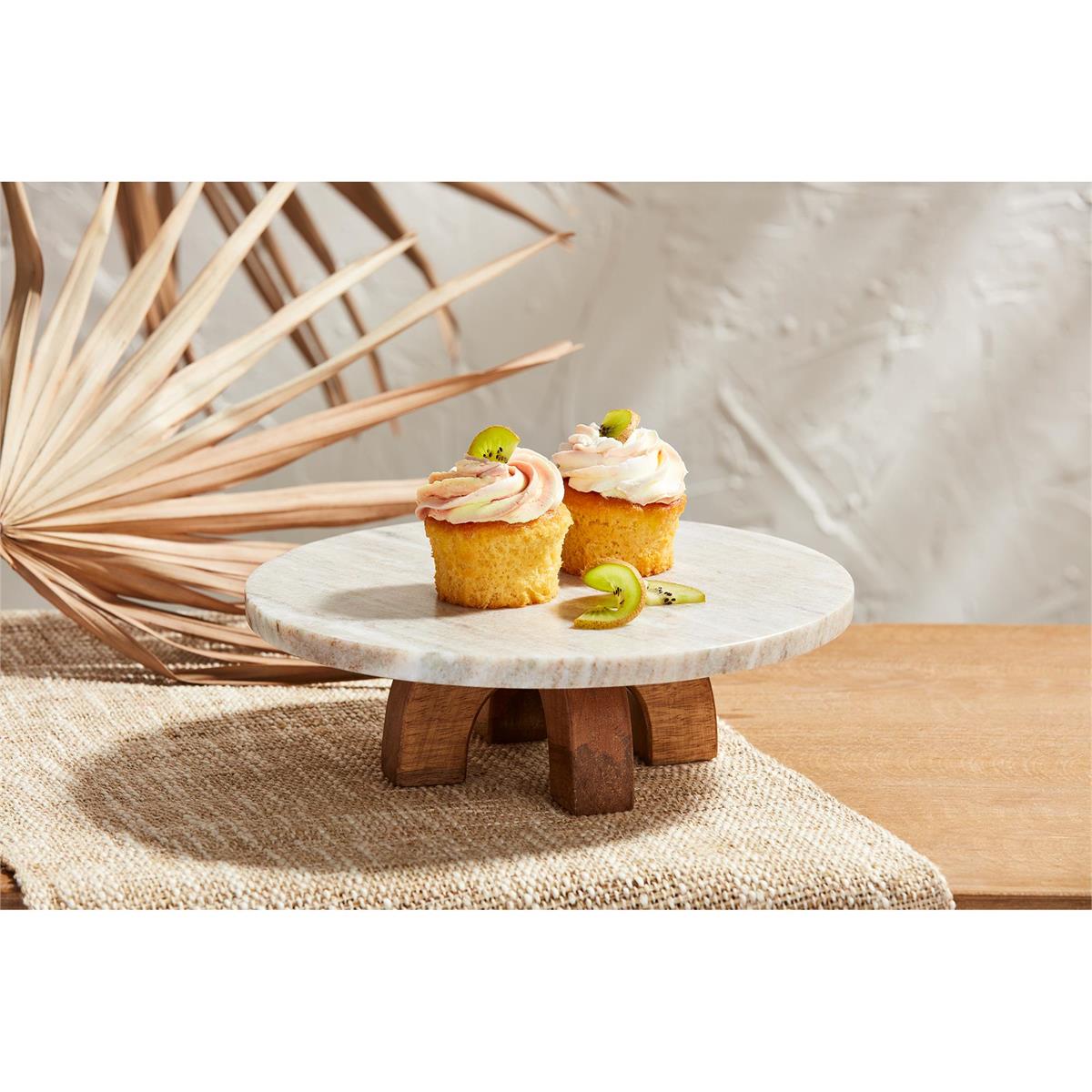 round marble slab on arched wooden legs with cupcakes on it set on a table with a brown linen table runner.