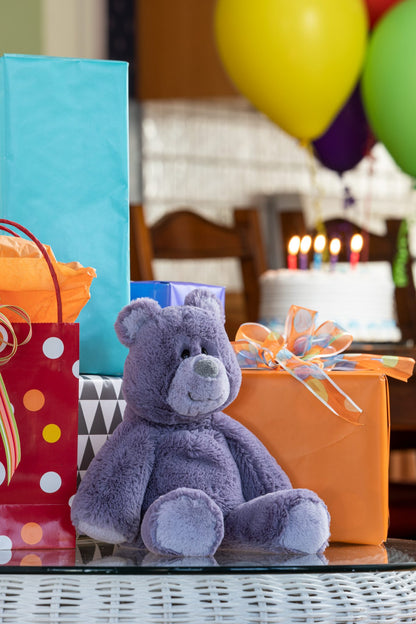Purple teddy bear with gift bags and balloons in the background