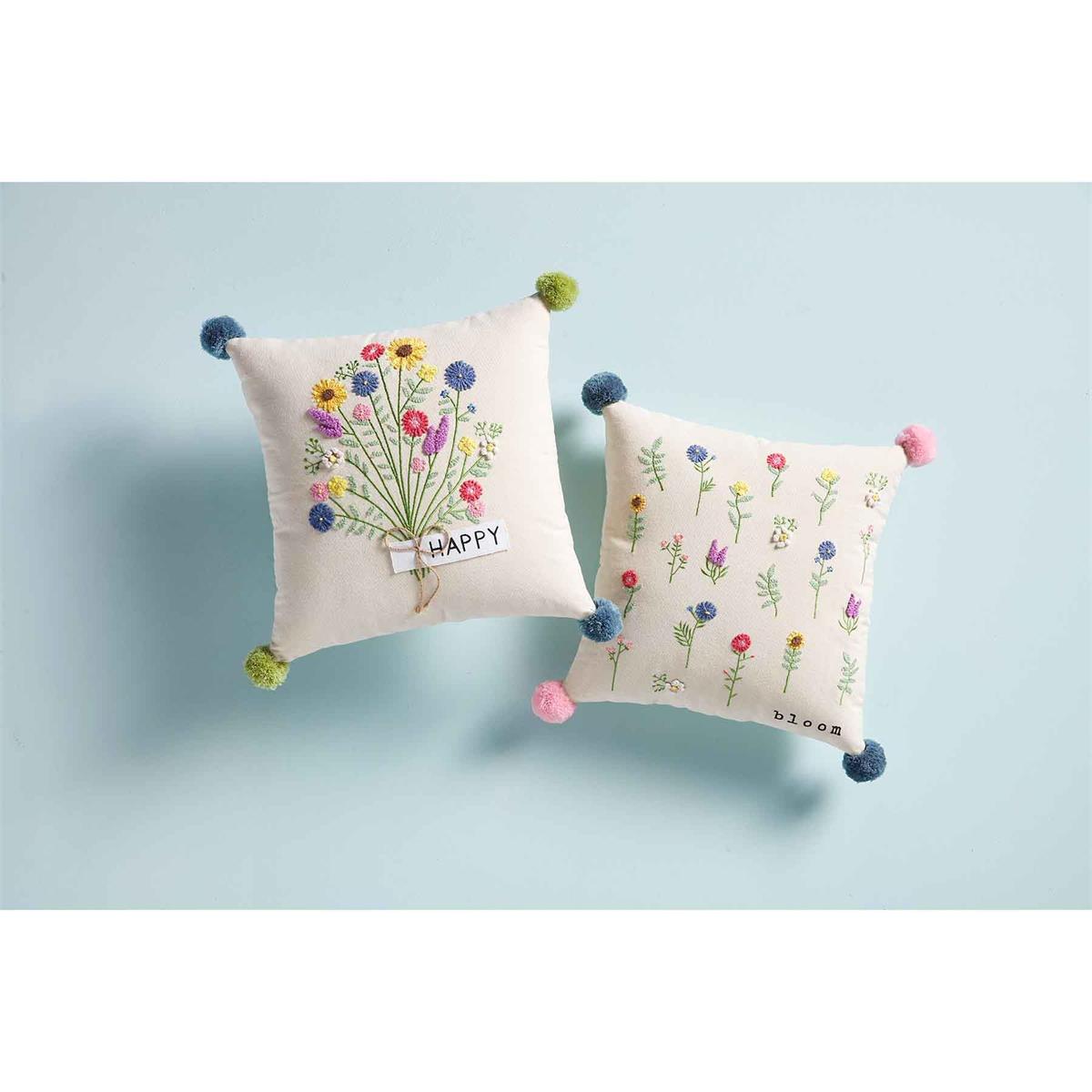 two floral embroidery pillows, one with happy and a spring floral bouquet and green and blue poms on the corners, the other has bloom and multiple colors of flowers scattered all over with pink and blue poms. both displayed against a light blue background