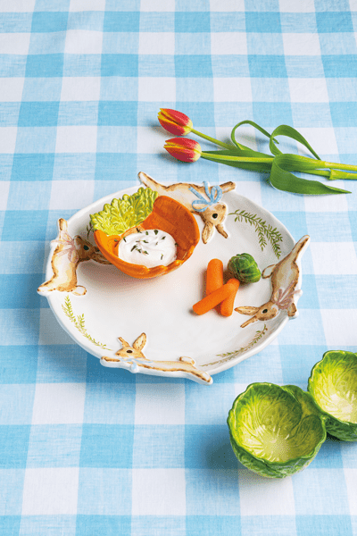 Decorative bowl with rabbit design and a smaller carrot bowl set inside it on a blue checkered tablecloth with vegetables and flowers.