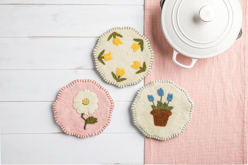 3 assorted Felted Floral Trivets set on a white wood surface with a pink cloth and a dutch oven