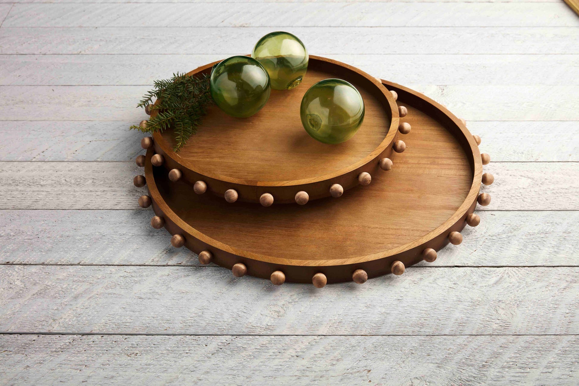 2 sizes of round wooden trays with beads around the rim on a grey slat table with greenery and orbs on them.