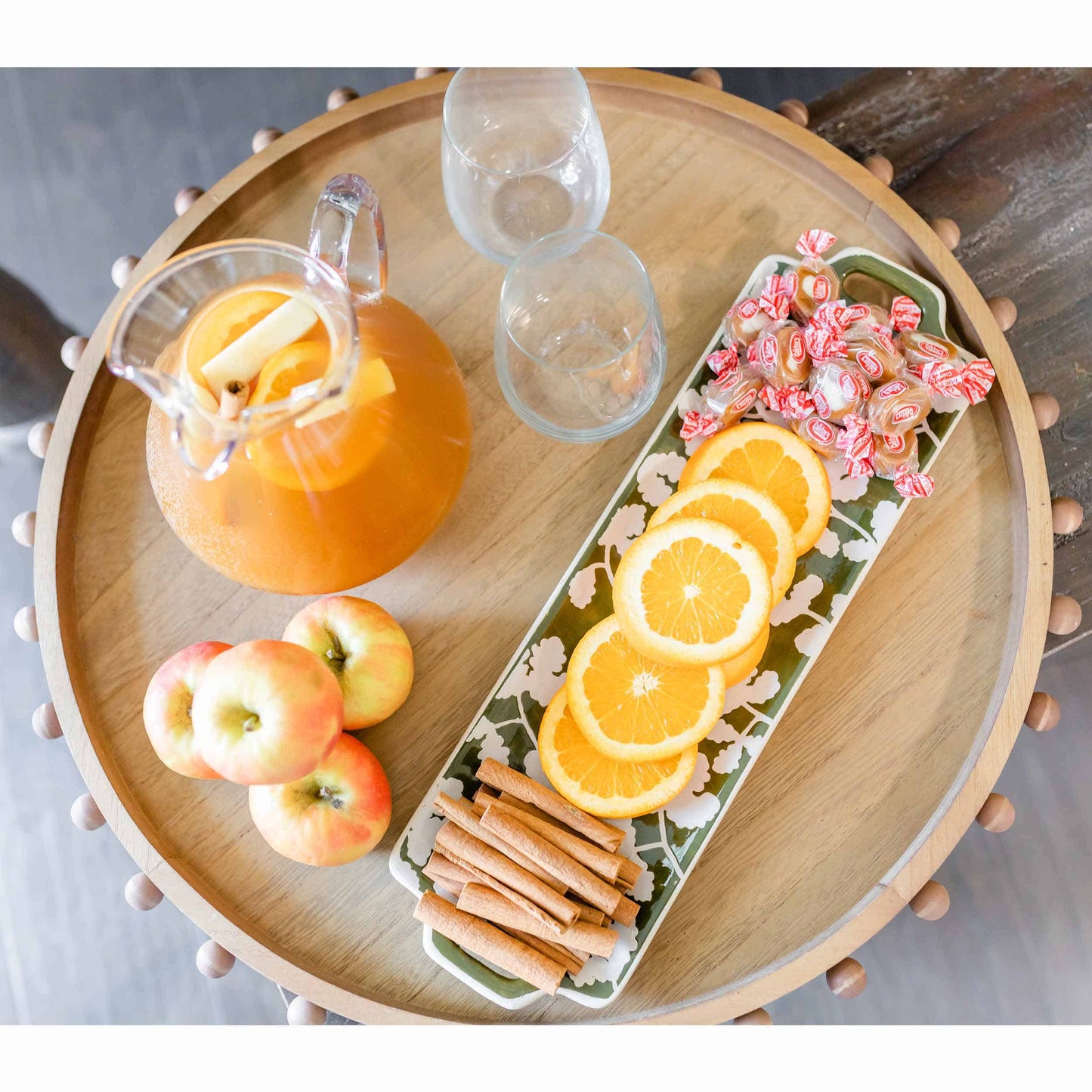 top view of round wooden tray on a table with a pitcher, glasses, apples and snacks arranged on it.