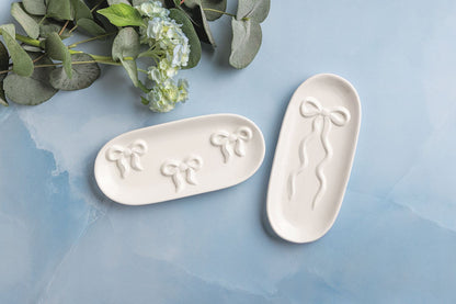 two oval trays with bow designs on them arranged on a blue surface with greenery to one side