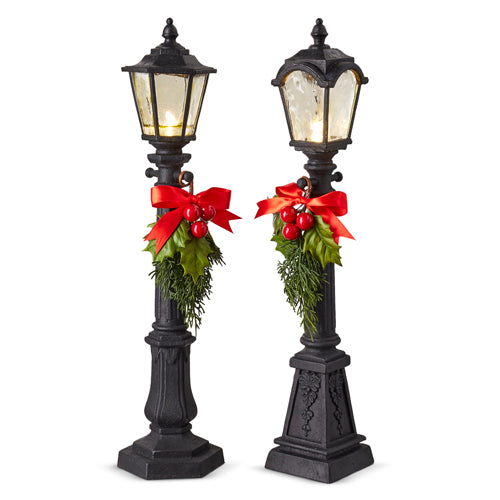 2 black lamps posts with greenery tied on them and the lights on.