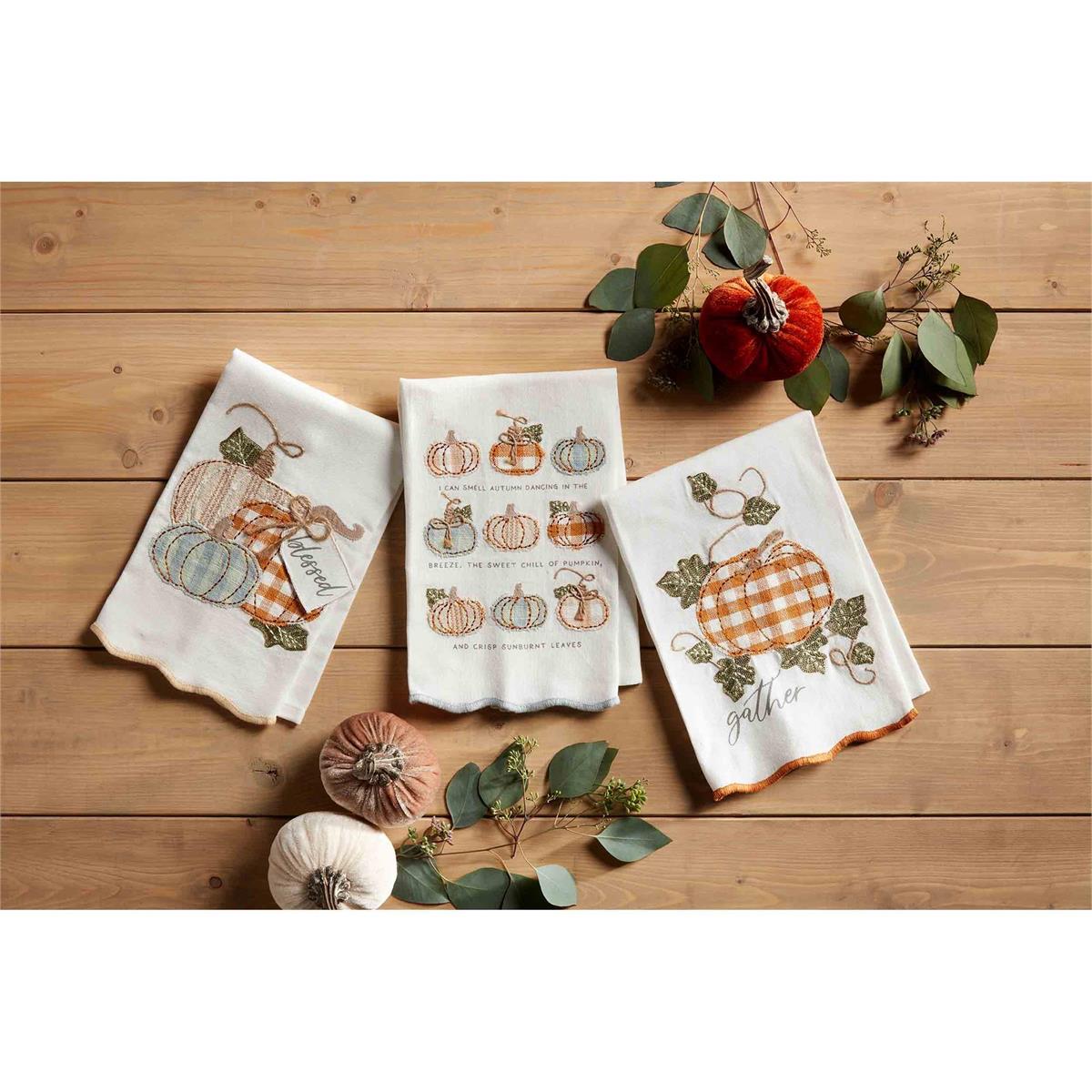 3 styles of appliqued fall towels arranged on a wood slat table with velvet pumpkins and greenery.