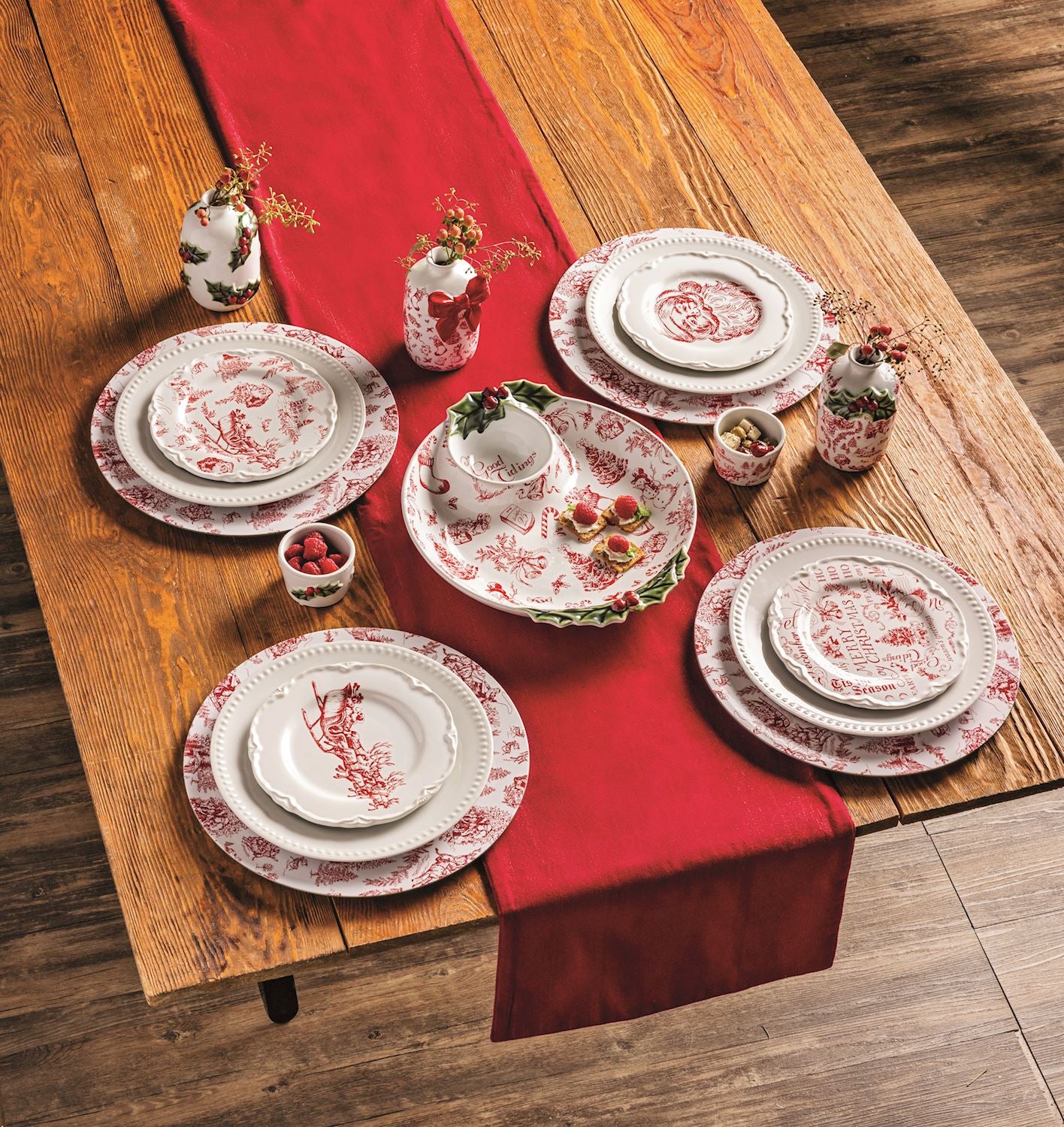 top view of a wooden table set with a red runner, toile chargers, plates, and other dishes.