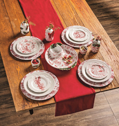 top view of a wooden table set with a red runner, toile chargers, plates, and other dishes.