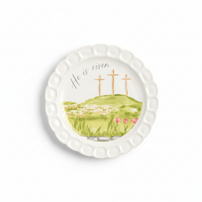 round white plate with hilly scene, 3 crosses, and he is risen printed on it