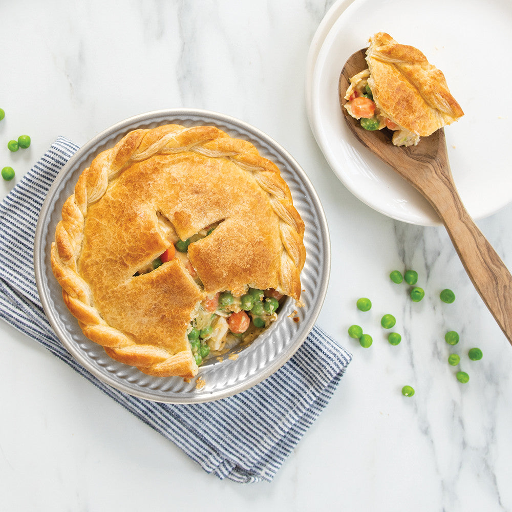 Personal Pie Pan filled with pot pie set on a dishtowel on a marble surface with a plate and wooden spoon and peas scattered about.