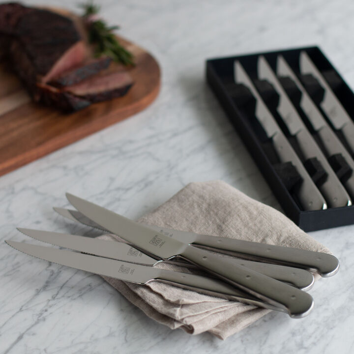 4 silver knives laying on a stack of linen napkins with 4 more knives in a box and a steak on a board in the background.