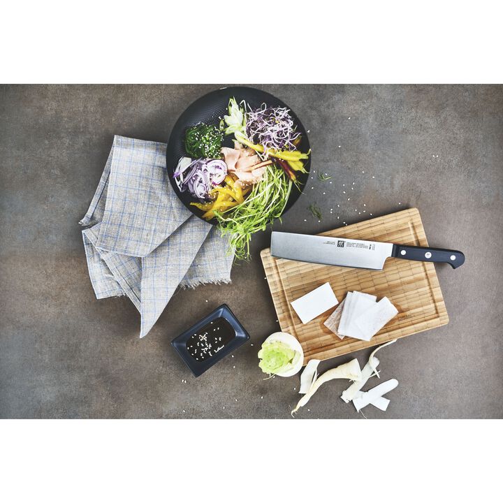 top view of countertop with Nakiri Knife laying on a cutting board surrounded with a bowl full of sliced veggies, a dish of herbs, ans a dish towel.